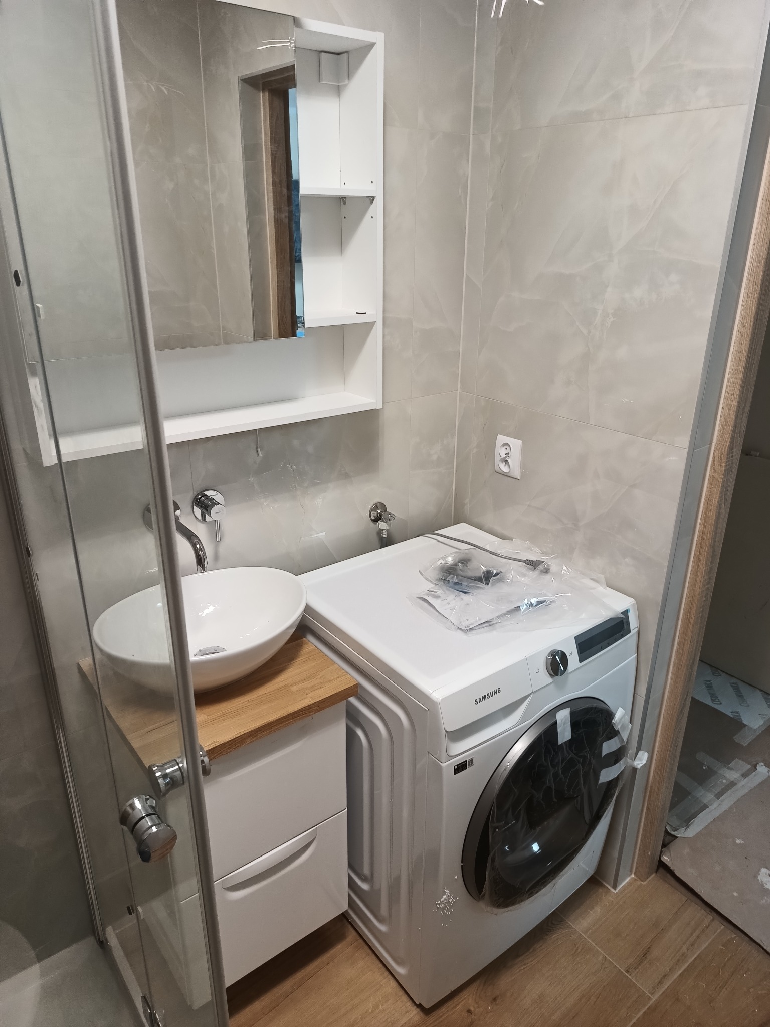 Nowoczesna łazienka z umywalką nablatową na drewnianym blacie, szafką pod umywalką, pralką Samsung z folią ochronną na drzwiczkach, kabiną prysznicową i lustrem z półkami.