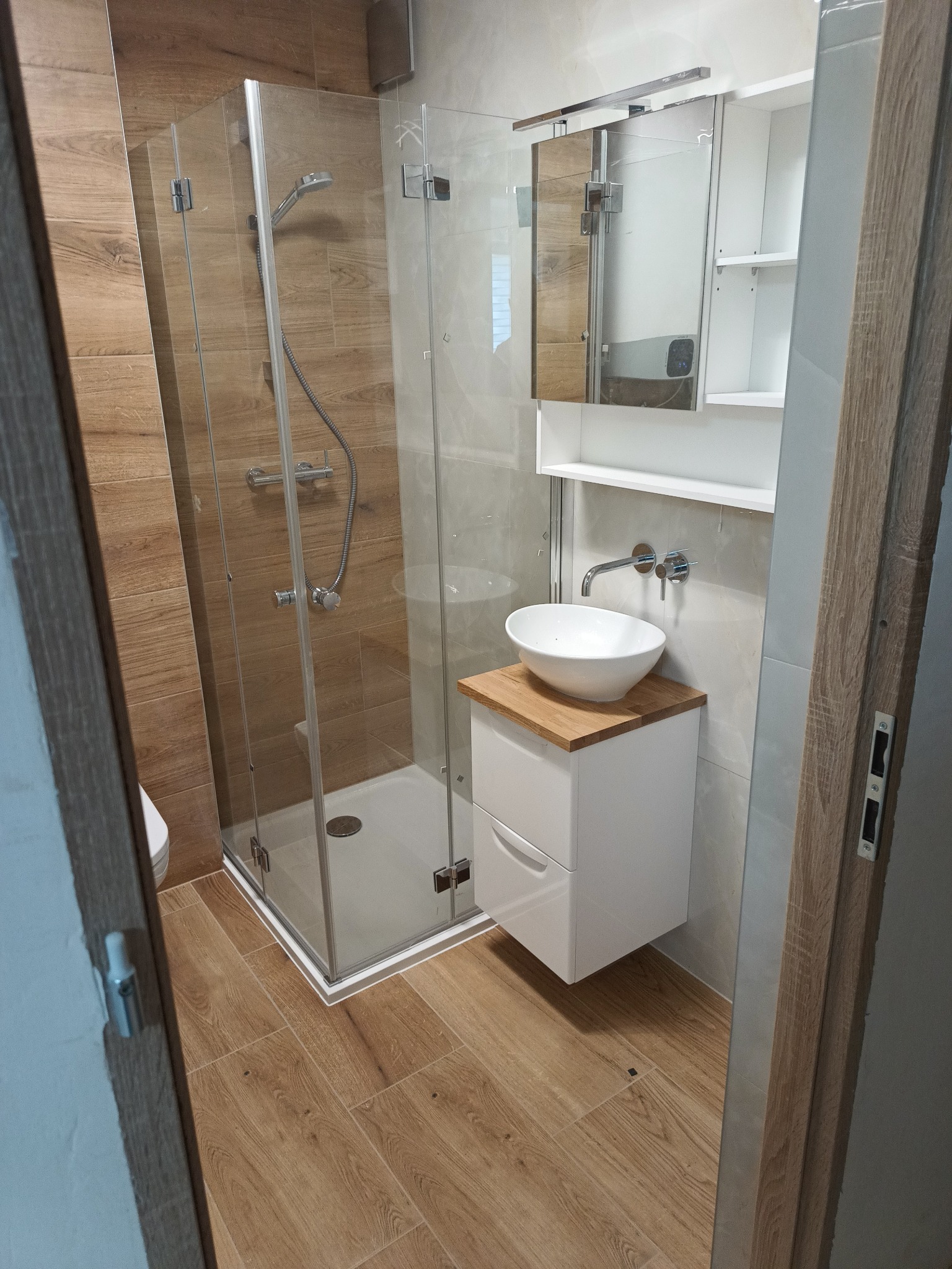 Nowoczesna łazienka w Puławach z kabiną prysznicową typu walk-in, białą umywalką nablatową na drewnianym blacie i lustrem z szafką, wykończona płytkami imitującymi drewno.