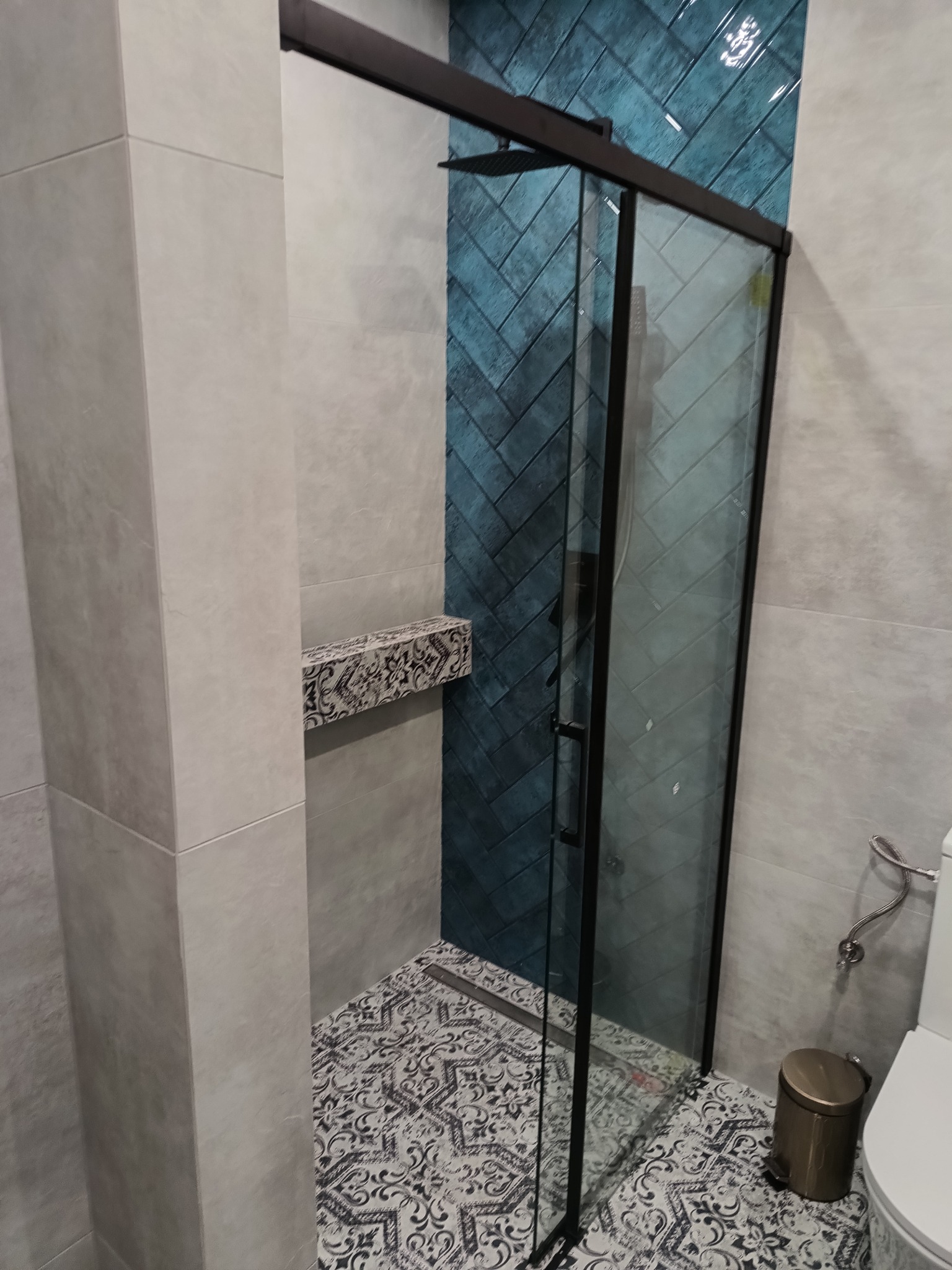 Nowoczesna kabina prysznicowa z czarnymi profilami, niebieskie płytki ułożone w jodełkę na ścianie wewnątrz kabiny, wzorzysta podłoga, szare płytki na pozostałych ścianach, półka z dekoracyjnym...