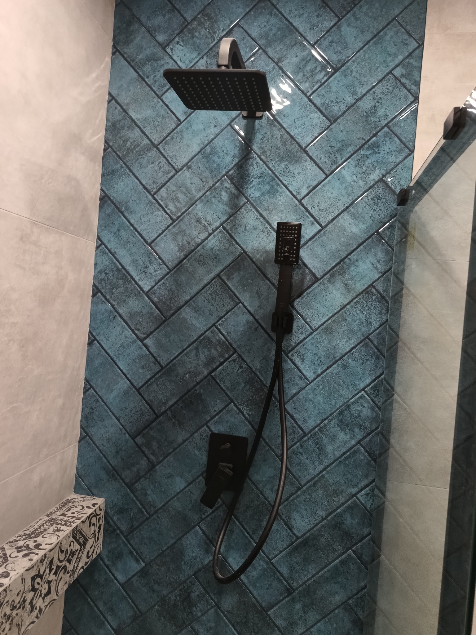 Ściana kabiny prysznicowej wyłożona turkusowymi, błyszczącymi płytkami ułożonymi w jodełkę, z czarną armaturą: deszczownicą, słuchawką prysznicową i baterią.