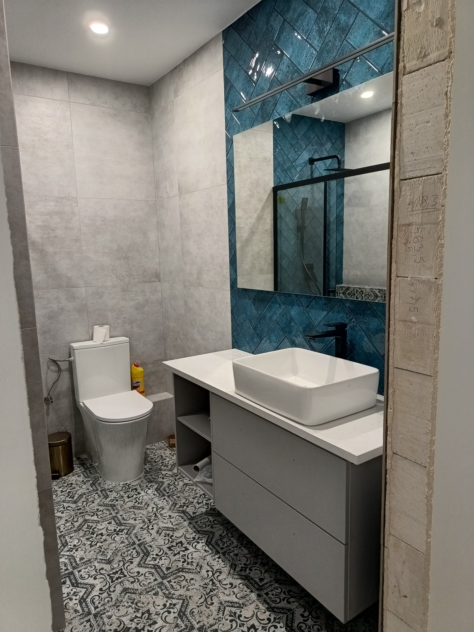 Nowoczesna łazienka z niebieskimi płytkami ułożonymi w jodełkę, szarą szafką pod umywalką i wzorzystą podłogą. W lustrze odbija się czarna kabina prysznicowa.