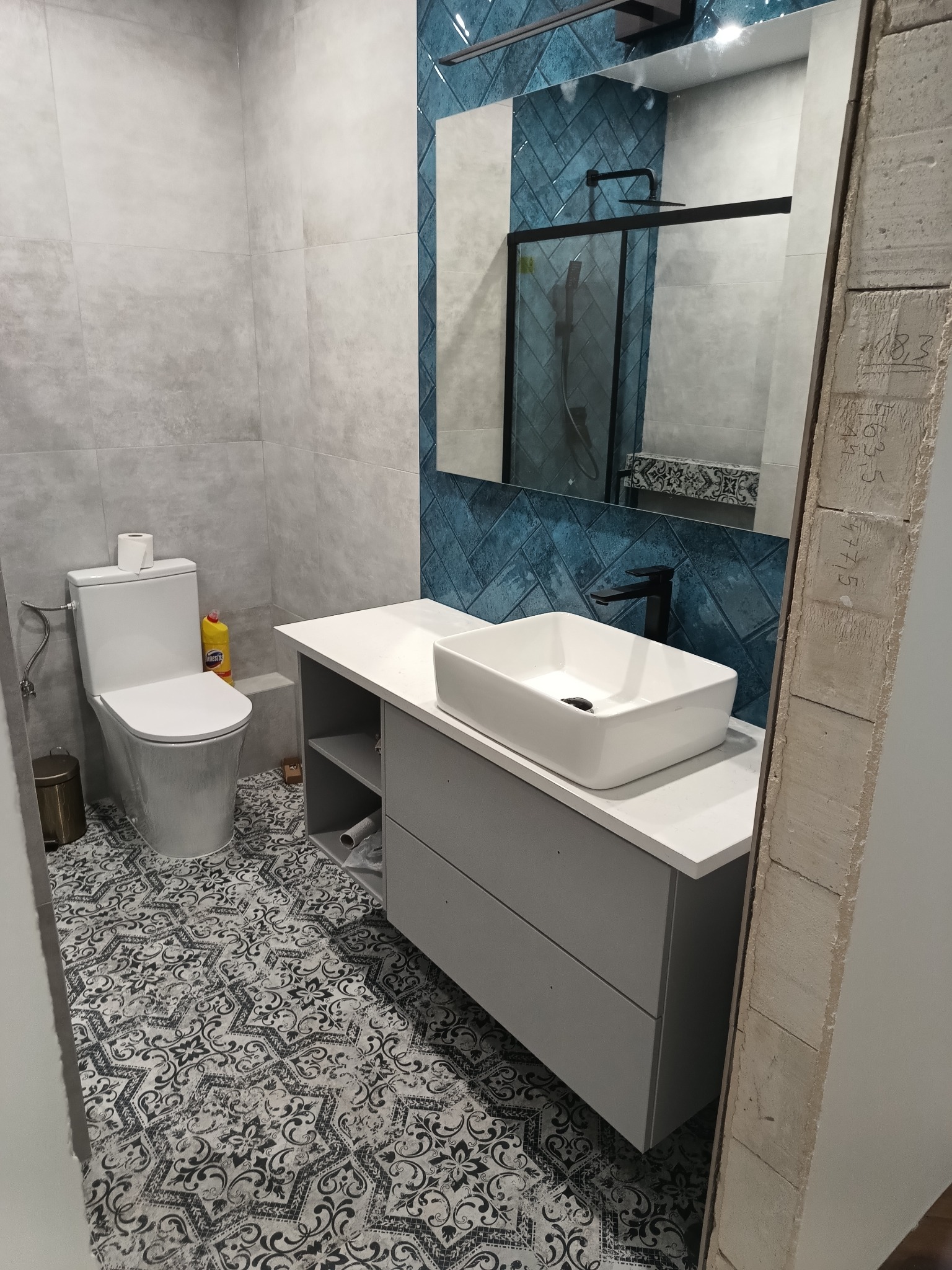 Nowoczesna łazienka z białą umywalką nablatową, szarą szafką z szufladami, toaletą, lustrem z niebieskimi płytkami ułożonymi w jodełkę i wzorzystą podłogą; widoczna butelka płynu do czyszczenia...