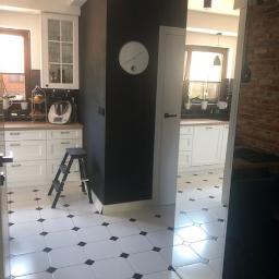 Benita Home & Garden - Nowoczesna kuchnia z białymi szafkami, czarną ścianą, biało-czarną podłogą w karo, widok na drugą część kuchni przez drzwi.