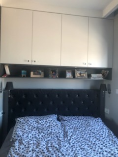 Sypialnia z ciemnym, pikowanym wezgłowiem łóżka i pościelą w drobny wzór. Nad łóżkiem szafki w kolorze białym i półka z dekoracjami, oświetlona lampkami nocnymi.