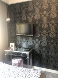 Czarno-srebrna tapeta w ornamenty na ścianie za czarną toaletką z lustrzanymi nogami i szarym pufem, nad toaletką zawieszony telewizor.