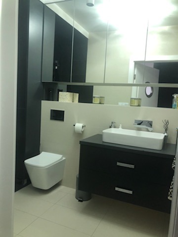 Nowoczesna łazienka z wiszącą toaletą, prostokątną umywalką nablatową na czarnej szafce z dwiema szufladami i lustrem na całej ścianie, odbijającym okrągłe okno w drzwiach.