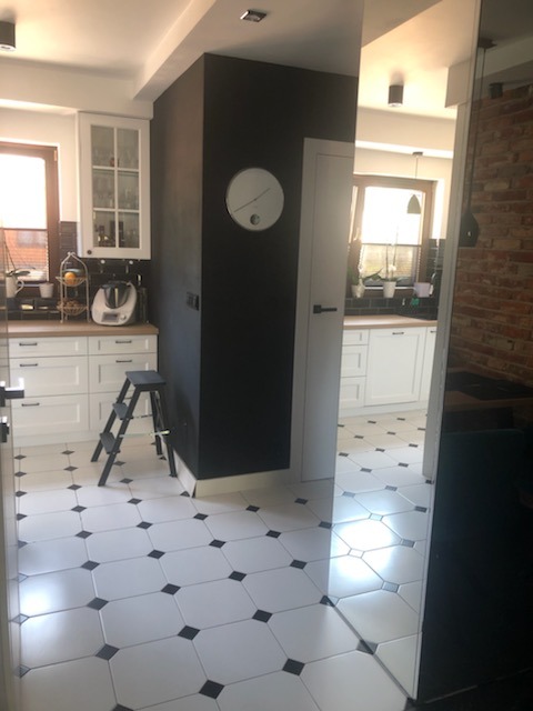Nowoczesna kuchnia z białymi szafkami, czarną ścianą, biało-czarną podłogą w karo, widok na drugą część kuchni przez drzwi.