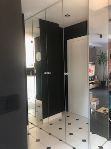 Wąski korytarz z lustrzanymi ścianami tworzącymi iluzję przestrzeni, biało-czarna podłoga w karo, widoczna część salonu z rośliną doniczkową w tle.