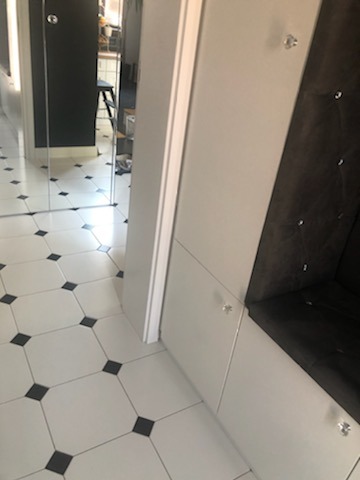 Wnęka z siedziskiem obita ciemną tkaniną z kryształkami, białe szafki z kryształowymi gałkami, podłoga z białych płytek z czarnymi wstawkami, lustro na ścianie odbijające wnętrze.