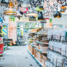 Wnętrze sklepu z artykułami do domu i ogrodu, z bogatą ekspozycją różnorodnych lamp wiszących pod sufitem, regałami wypełnionymi koszami wiklinowymi, ziemią do kwiatów i tekstyliami domowymi.