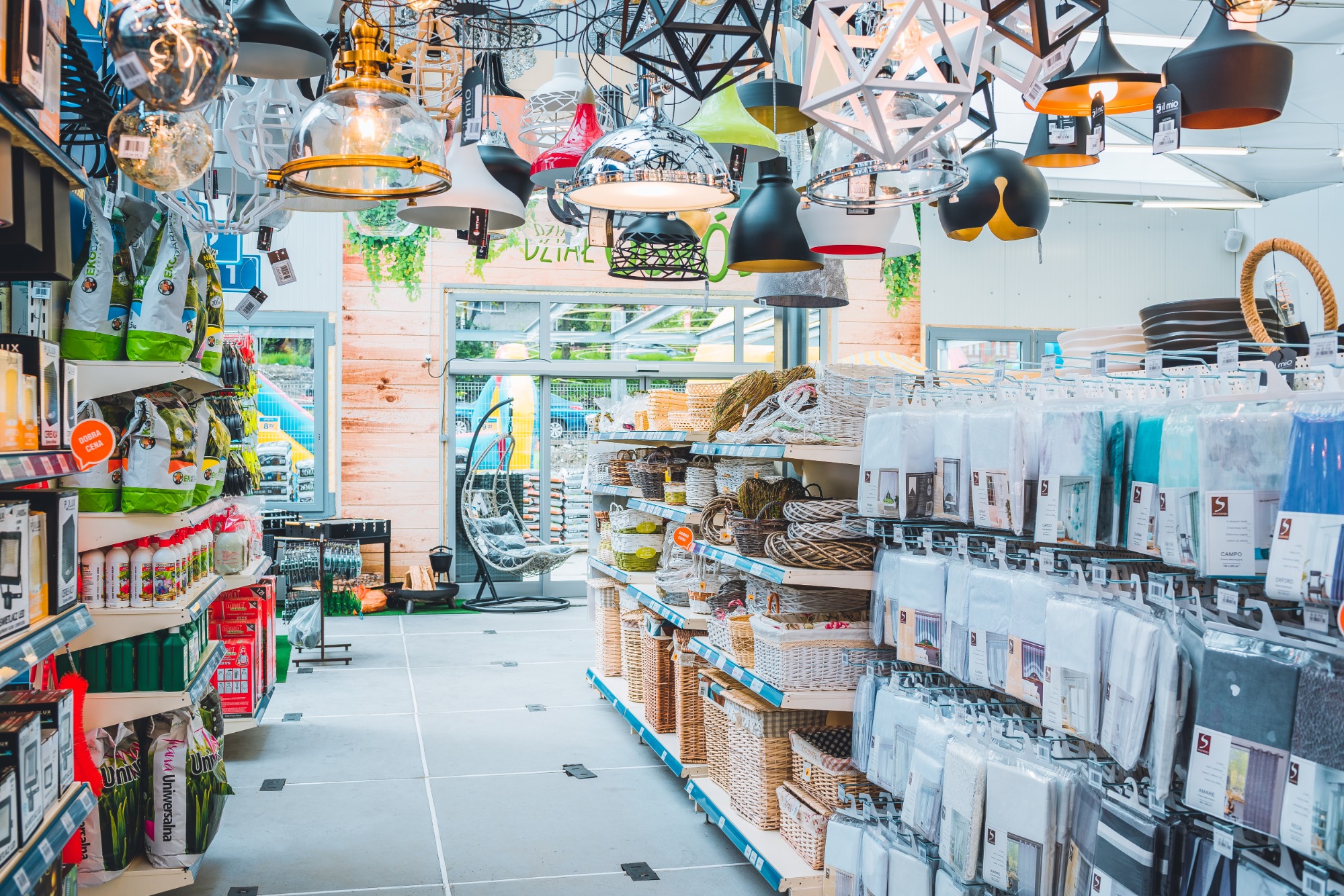 Wnętrze sklepu z artykułami do domu i ogrodu, z bogatą ekspozycją różnorodnych lamp wiszących pod sufitem, regałami wypełnionymi koszami wiklinowymi, ziemią do kwiatów i tekstyliami domowymi.