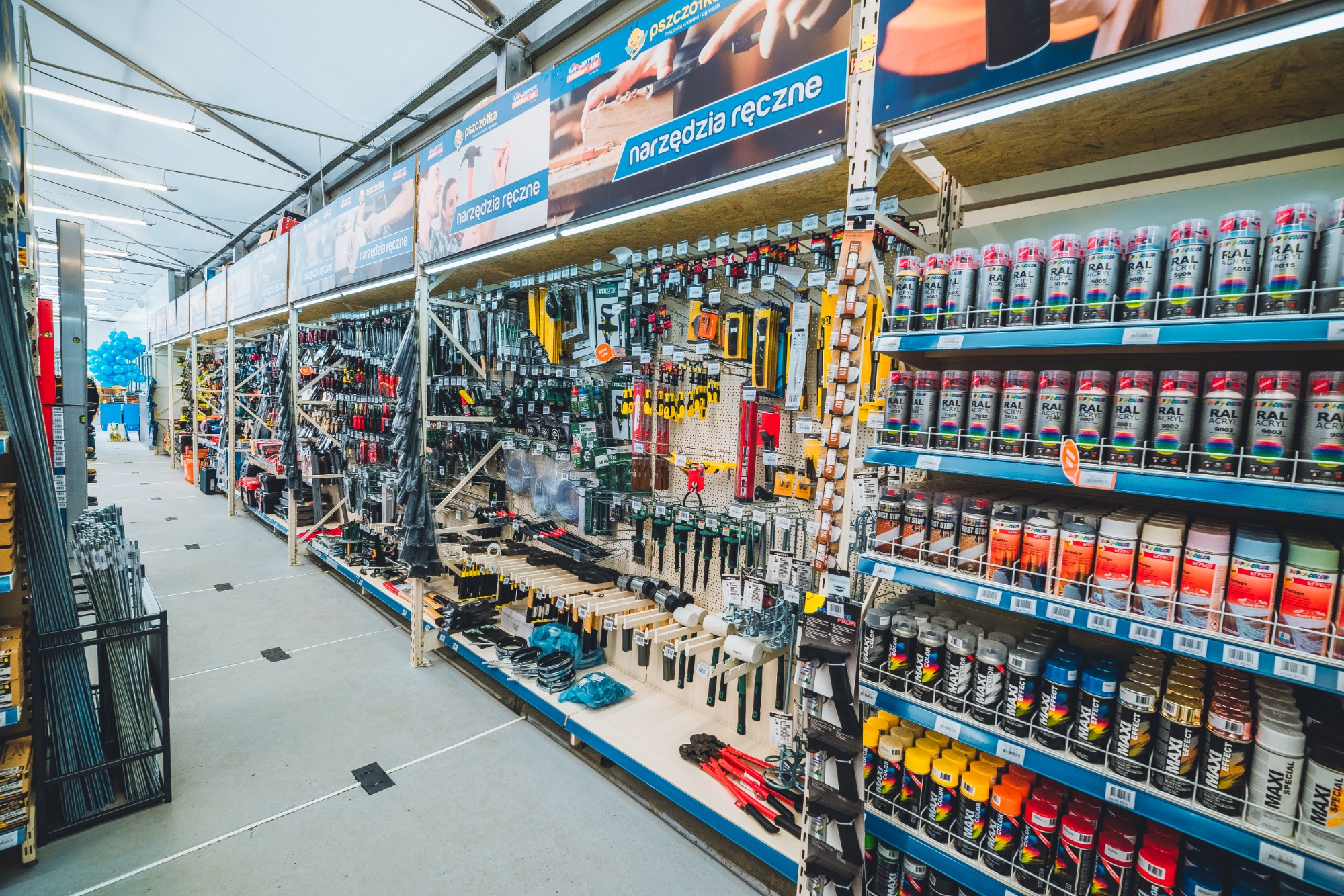 Wnętrze sklepu budowlanego z szerokim asortymentem narzędzi ręcznych i farb w sprayu na półkach, widok wzdłuż alejki między regałami.