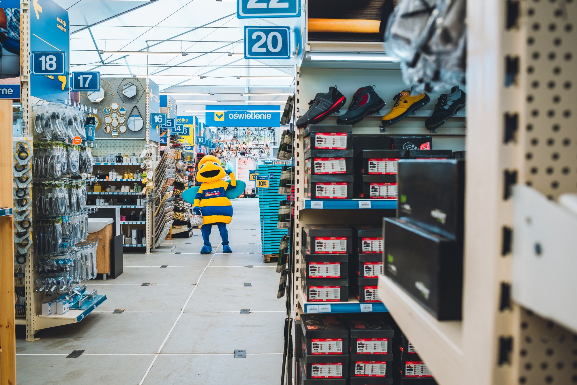 Wnętrze sklepu budowlanego z oświetleniem i artykułami sanitarnymi, w tle maskotka w stroju pszczoły z koszykiem w ręku.