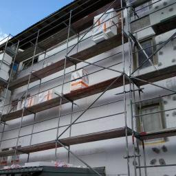 Zorza-Budownictwo - Ocieplanie budynku styropianem, widoczne rusztowania i paczki styropianu marki KRASBUD na różnych poziomach, niektóre okna zabezpieczone folią.