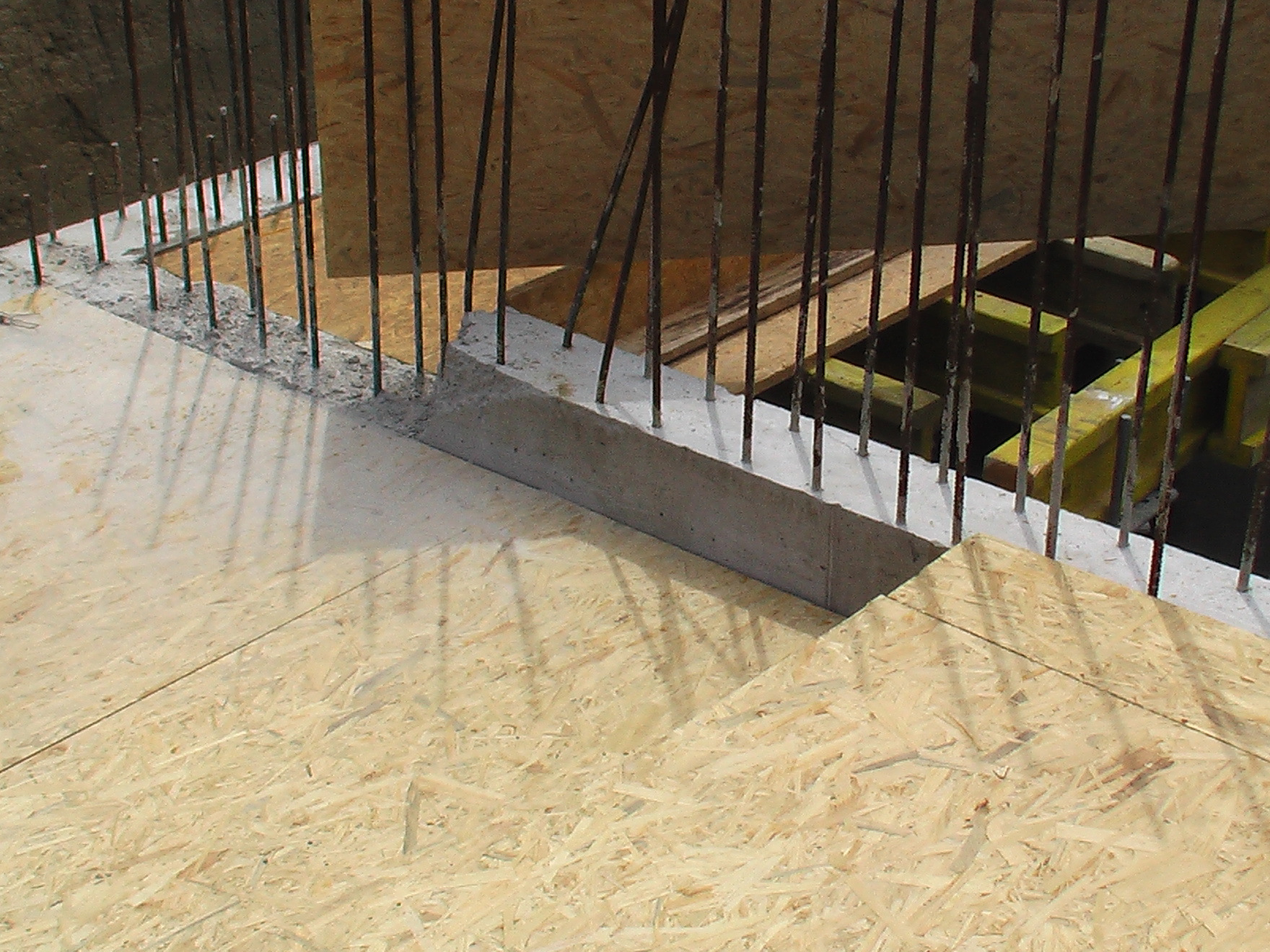 Szare, betonowe wzmocnienie z wystającymi prętami zbrojeniowymi, widoczne na tle drewnianego szalunku i żółtych elementów konstrukcyjnych.