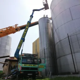 Niebieski podnośnik koszowy z numerem telefonu na wysięgniku, ustawiony przy dużych, srebrnych silosach przemysłowych. W koszu podnośnika znajduje się osoba pracująca na wysokości.
