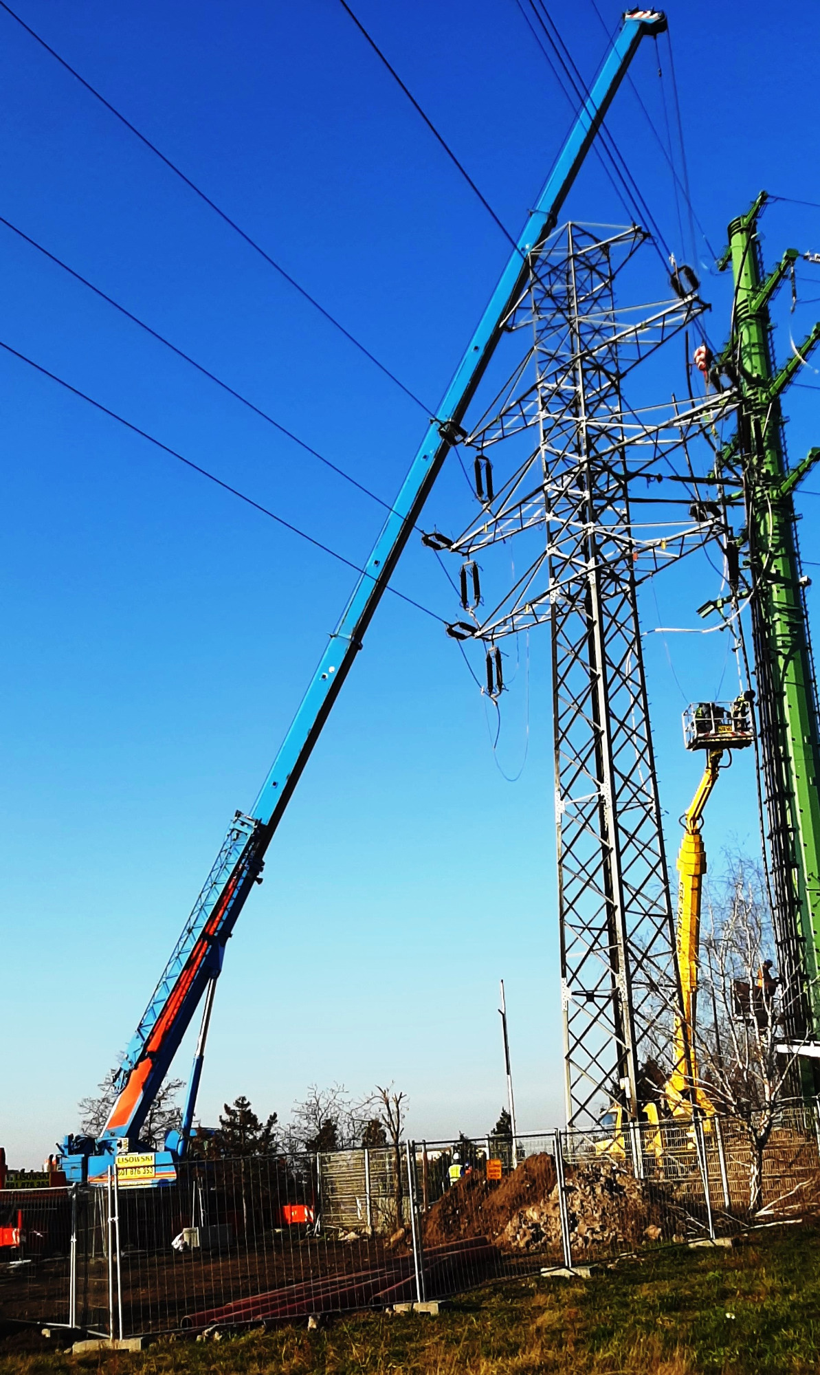 Prace konserwacyjne na słupach wysokiego napięcia z użyciem niebieskiego dźwigu z pomarańczowymi akcentami i żółtego podnośnika koszowego na tle błękitnego nieba.