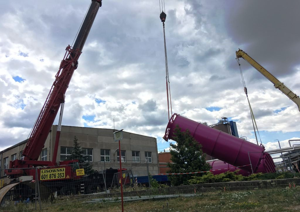 Dwa dźwigi, czerwony i żółty, unoszą duży, purpurowy zbiornik na tle pochmurnego nieba i przemysłowego budynku.