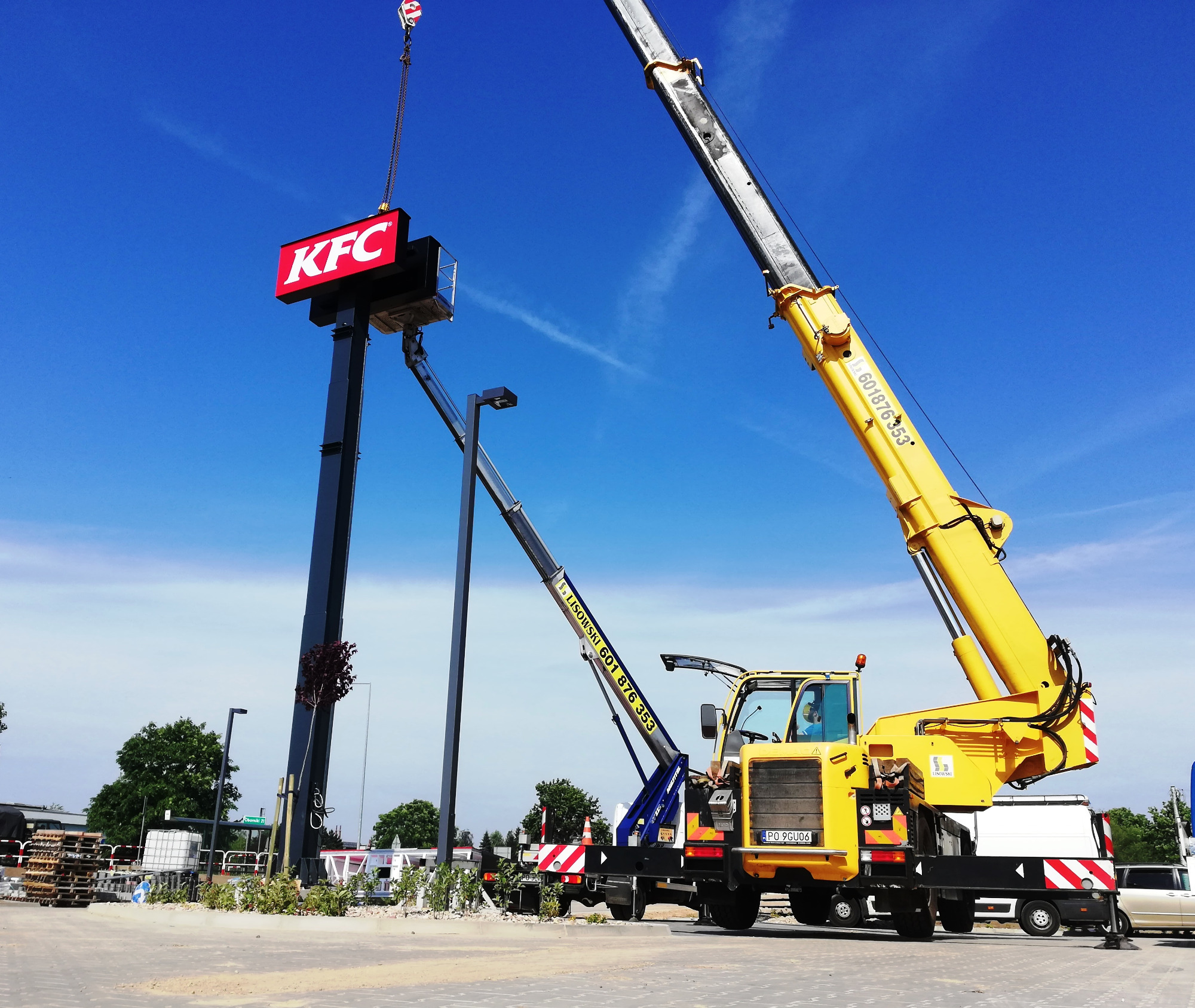 Montaż podświetlanego szyldu KFC za pomocą dwóch dźwigów na parkingu, widoczne numery telefonów na ramionach dźwigów, błękitne niebo.
