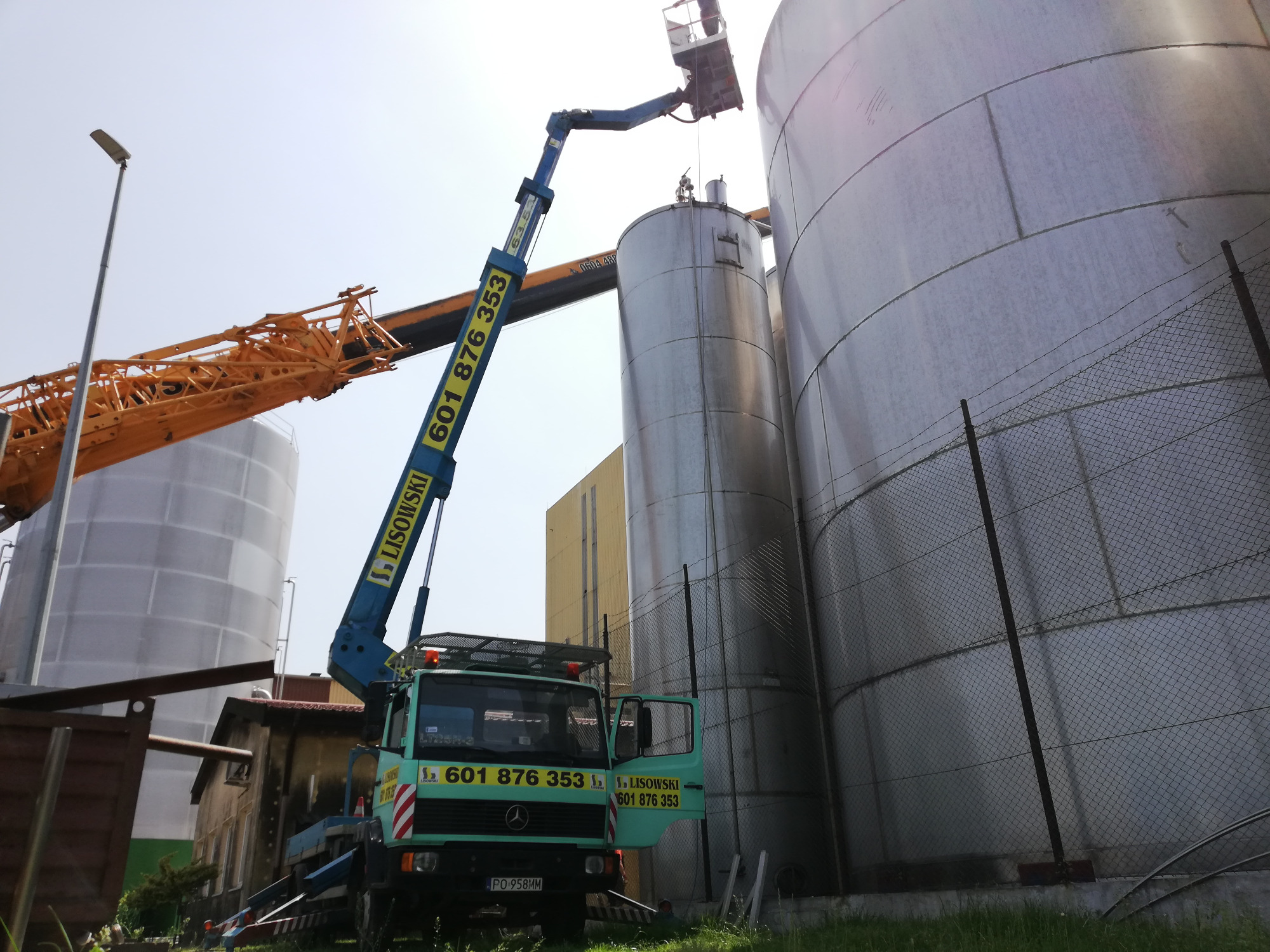 Niebieski podnośnik koszowy z numerem telefonu na wysięgniku, ustawiony przy dużych, srebrnych silosach przemysłowych. W koszu podnośnika znajduje się osoba pracująca na wysokości.