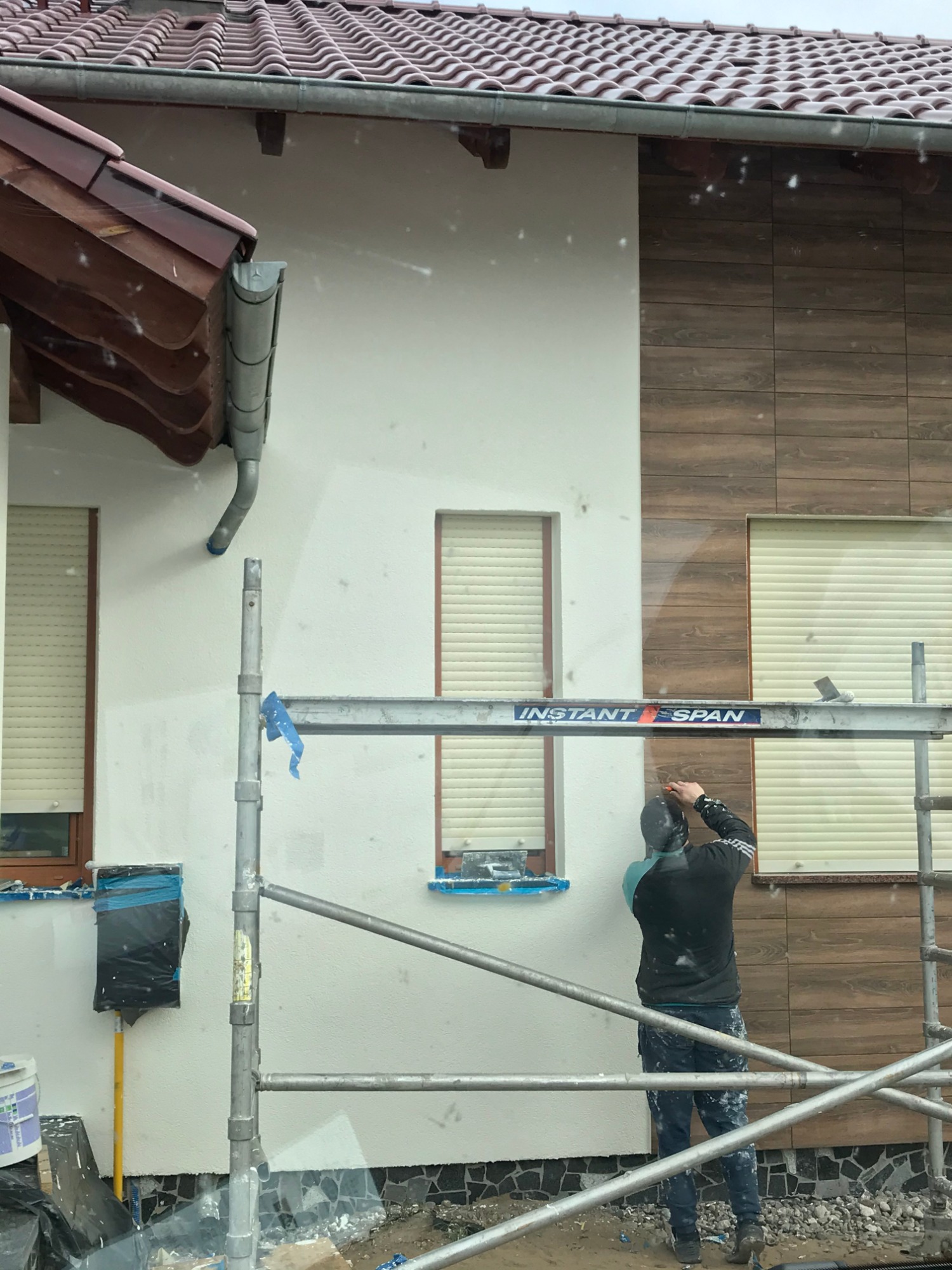 Pracownik na rusztowaniu montuje płytki elewacyjne imitujące drewno na fragmencie ściany budynku jednorodzinnego, obok widoczna pomalowana na biało ściana i okna z roletami.