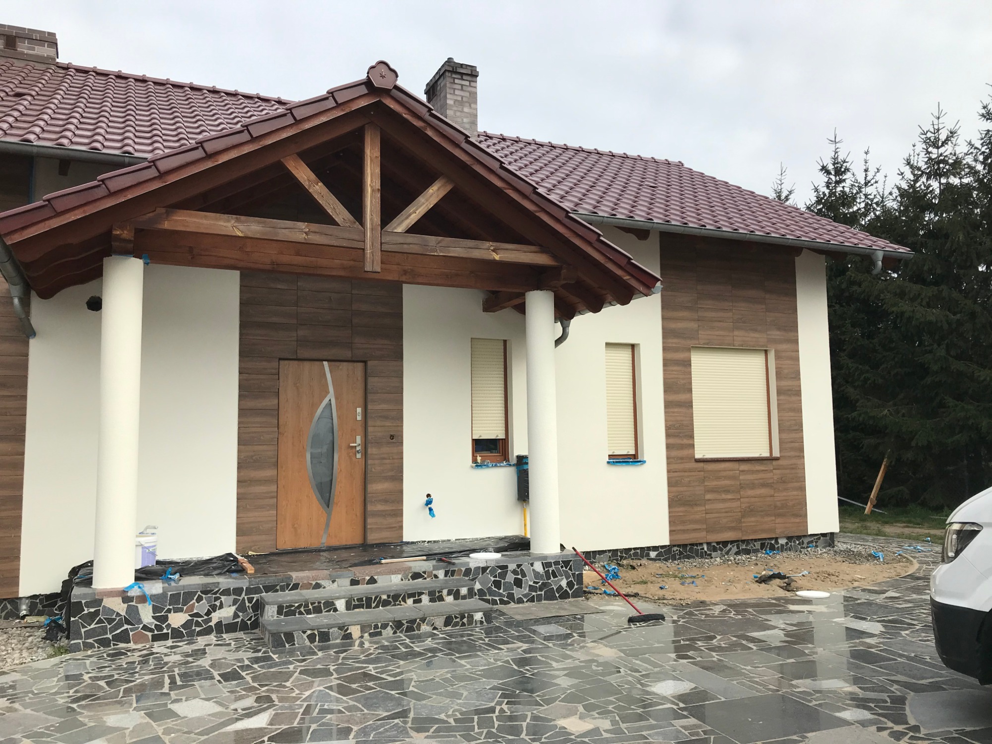 Nowoczesny dom jednorodzinny z jasną elewacją, drewnianymi elementami dekoracyjnymi i brukowanym podjazdem, widoczne ślady po pracach wykończeniowych wokół budynku.