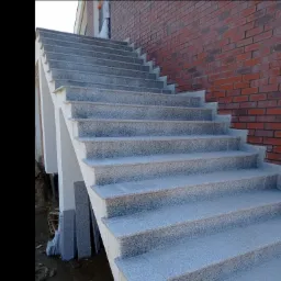 Zewnętrzne schody pokryte szarą, nakrapianą płytą kamienną, prowadzące do budynku z ceglaną ścianą. U dołu stos płytek gotowych do montażu.