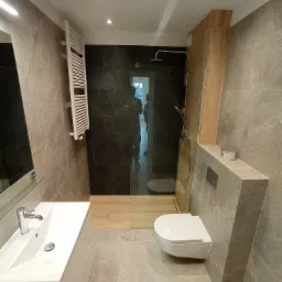 Nowoczesna łazienka z szarymi płytkami imitującymi marmur, kabiną prysznicową typu walk-in z czarną ścianą, podwieszaną toaletą, białą umywalką z szafką i grzejnikiem drabinkowym.