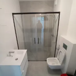 Nowoczesna łazienka z kabiną prysznicową typu walk-in z czarną ramą, białą umywalką z szafką, podwieszaną toaletą i szarą płytką na ścianie oraz podłodze. W lustrze kabiny odbija się sylwetka osoby.