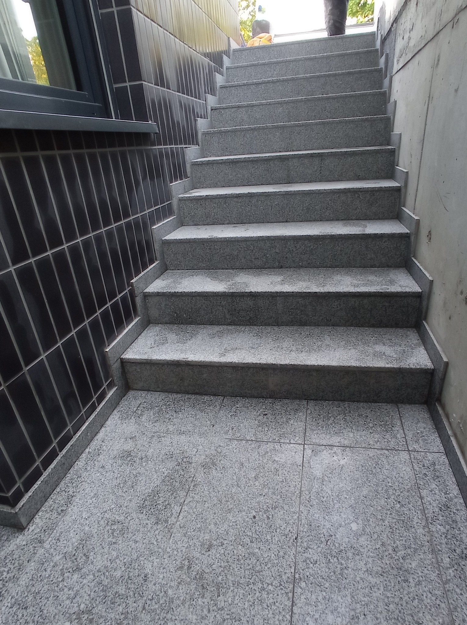 Granitowe schody zewnętrzne z wykończeniem z ciemnych płytek ceramicznych, widoczne z dołu, z fragmentem osoby w górnej części kadru.