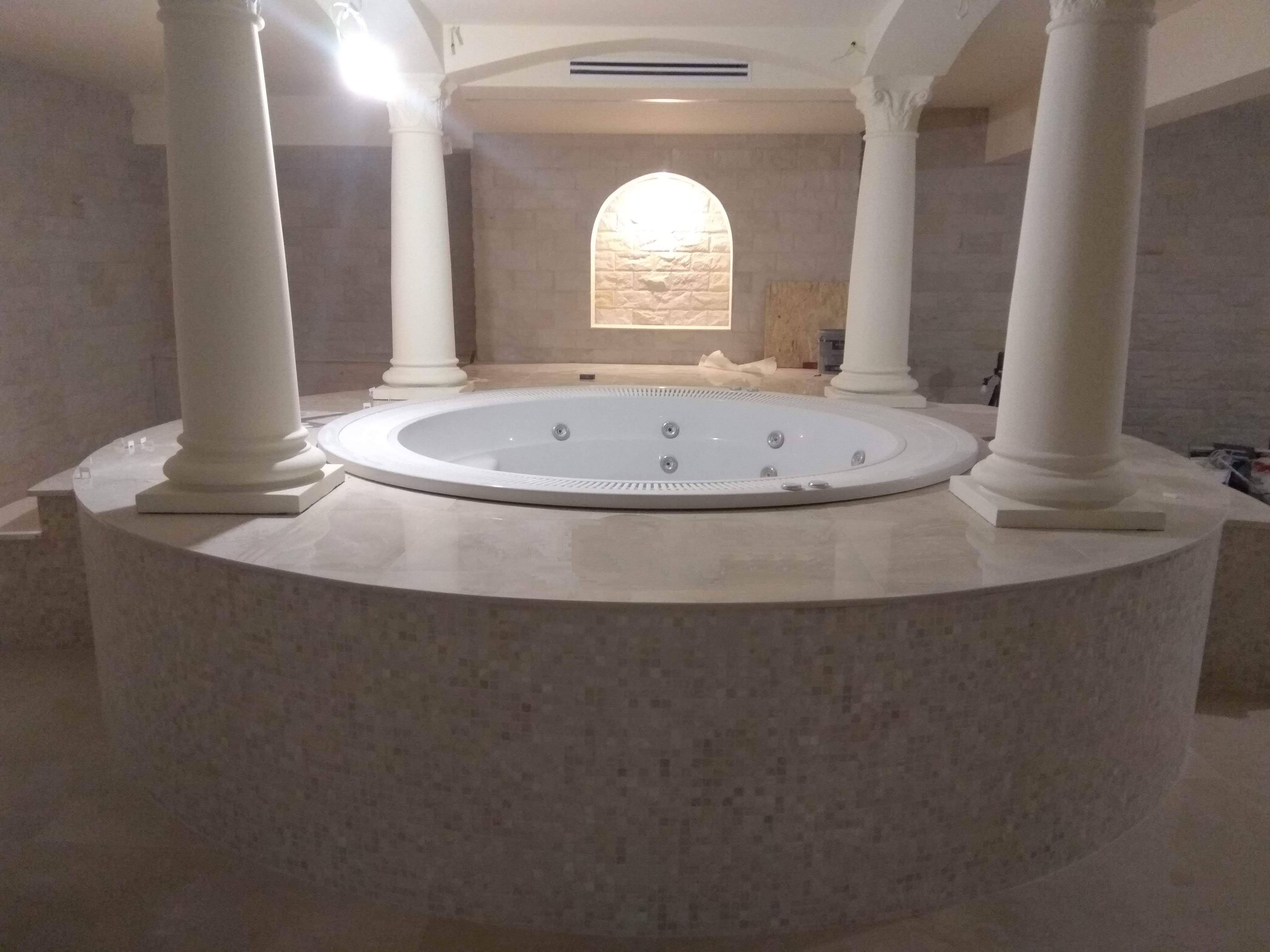 Okrągła wanna z hydromasażem na podwyższeniu z mozaiki w łazience z kolumnami i oświetloną wnęką w ścianie z kamienia.