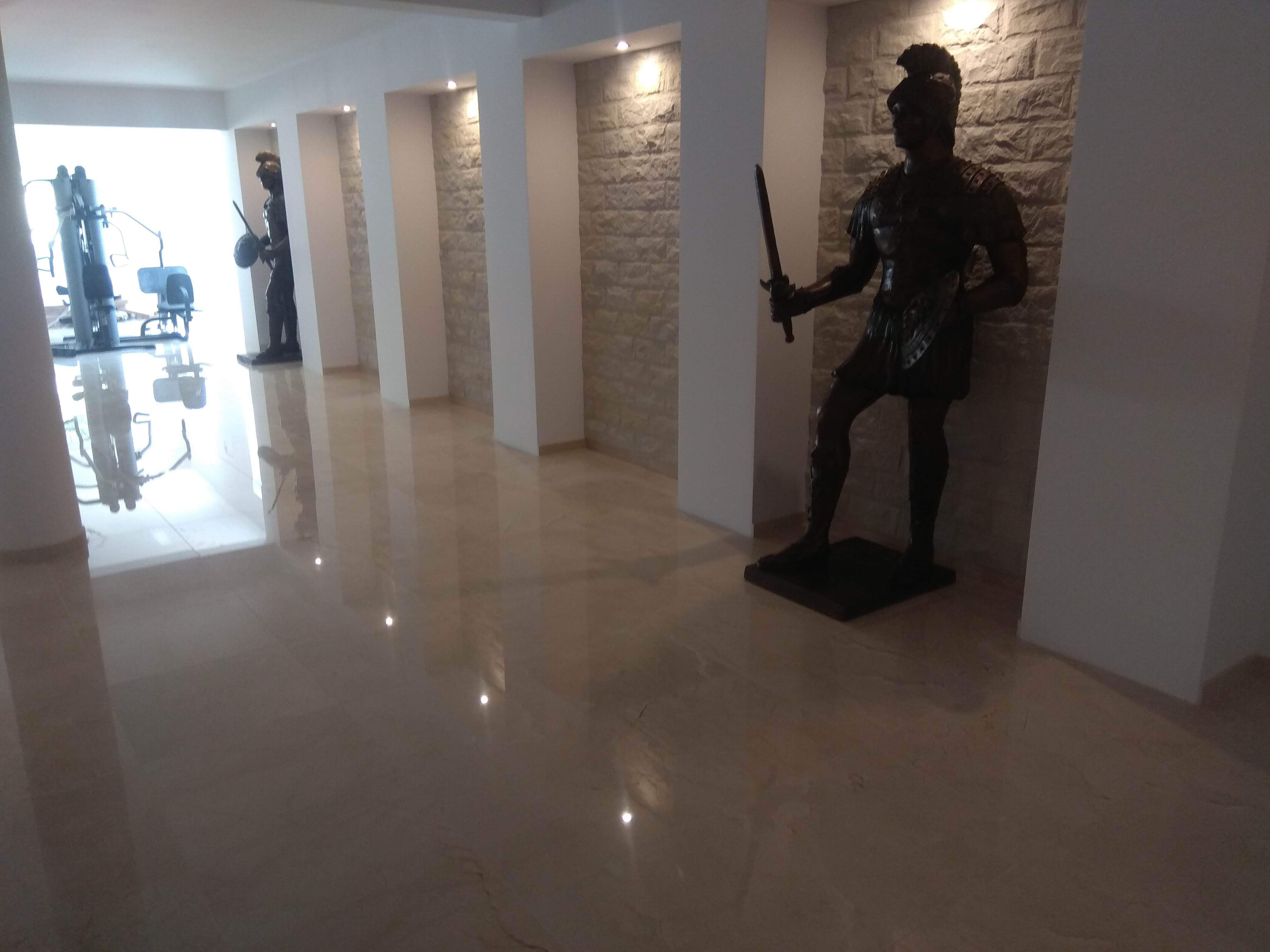 Polerowany beżowy marmur na podłodze korytarza, z wnękami w ścianach wyłożonymi kamieniem i rzeźbami rzymskich legionistów, oświetlone punktowymi lampami.