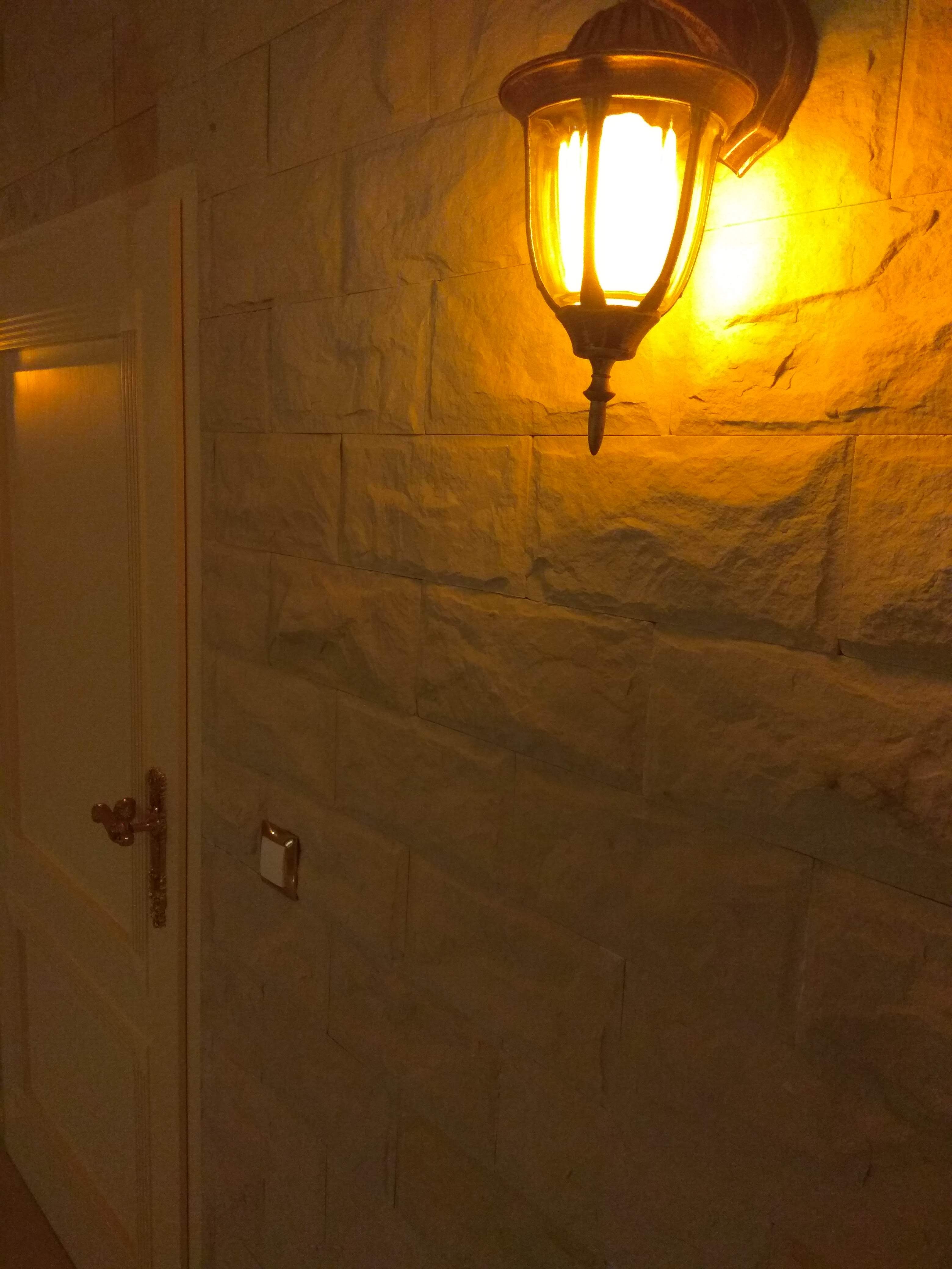 Ściana wyłożona jasną, nieregularną kamienną okładziną, oświetlona przez klasyczny kinkiet z ciepłym światłem, obok widoczny fragment drzwi z ozdobną klamką.