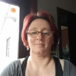 Portret kobiety w okularach z rudymi włosami, stojącej w pomieszczeniu z oknem ozdobionym naklejką w kształcie kwiatka i ciemnymi drzwiami w tle.