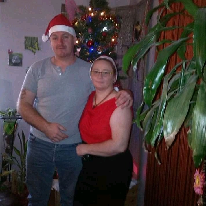 Para w świątecznych nakryciach głowy pozuje przed choinką i dużą rośliną doniczkową, mężczyzna obejmuje kobietę ramieniem.