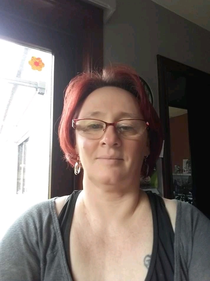 Portret kobiety w okularach z rudymi włosami, stojącej w pomieszczeniu z oknem ozdobionym naklejką w kształcie kwiatka i ciemnymi drzwiami w tle.