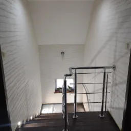 Schody w dół z ciemnobrązowymi stopniami, oświetleniem w podstopnicach i chromowaną balustradą. Ściany wykończone dekoracyjnym tynkiem w odcieniach beżu i bieli.