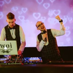 Dwóch wodzirejów w kamizelkach w kratę, jeden z mikrofonem, drugi obsługuje sprzęt DJski z laptopem z logo firmy na tle ekranu z motywem serc.
