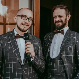 Dwóch mężczyzn w kratkowanych garniturach i drewnianych muchach, jeden z mikrofonem, uśmiechają się do kamery, drugi mruga.