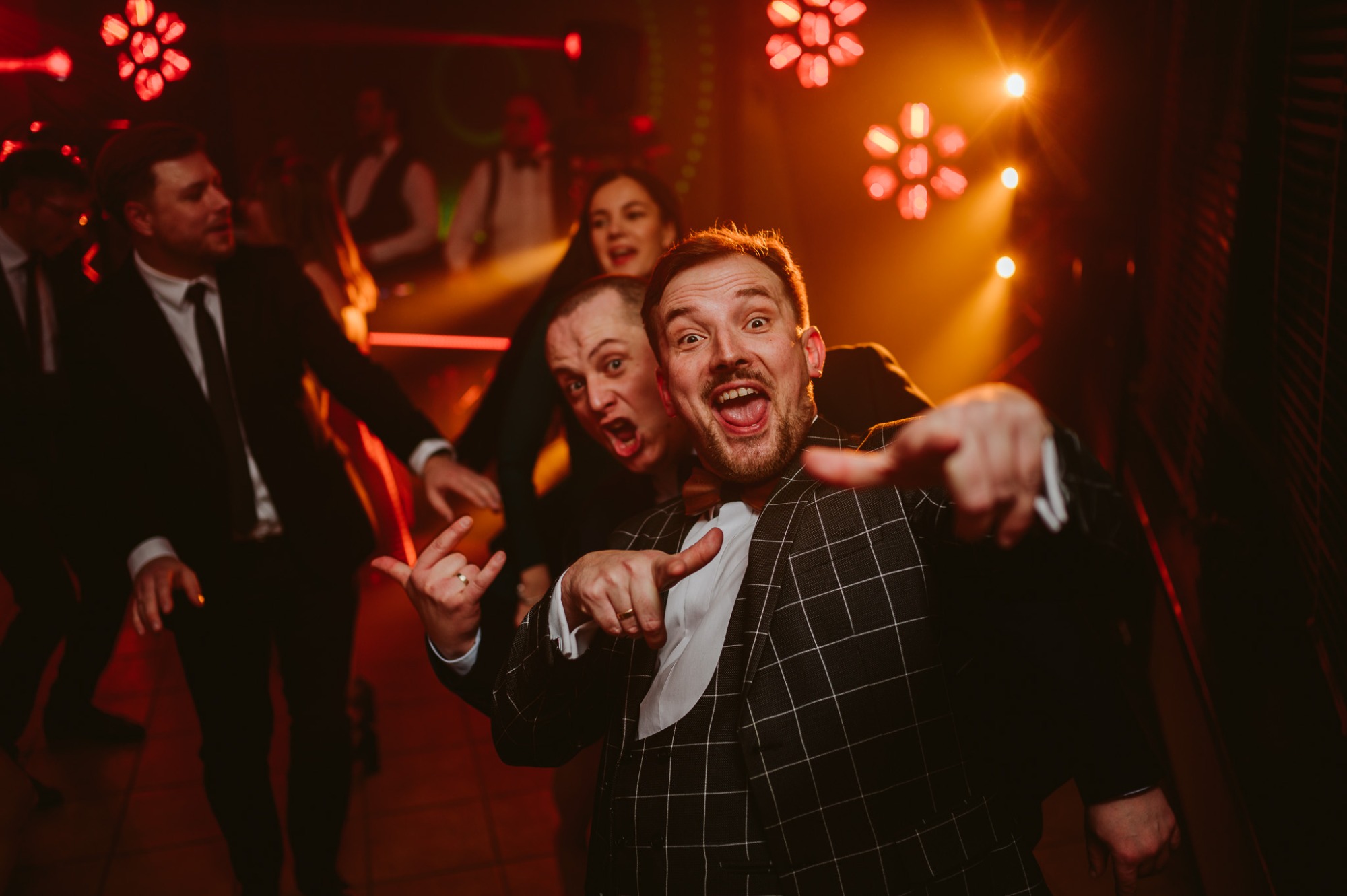 Wesoła grupa osób w garniturach, w tym mężczyzna w kraciastej marynarce z muszką, wskazujący palcem w stronę obiektywu na tle scenicznego oświetlenia i bawiących się ludzi.