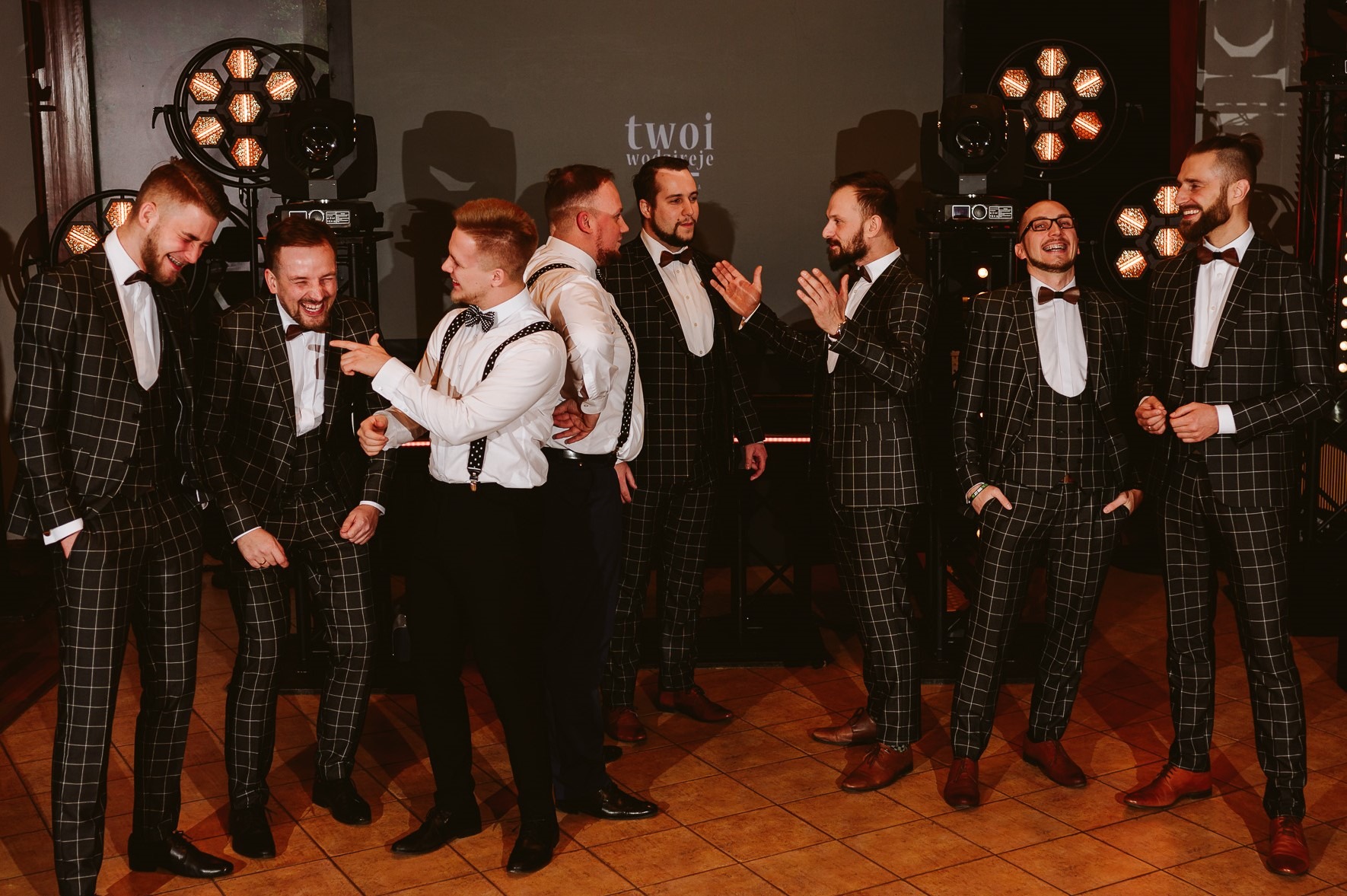 Grupa mężczyzn w eleganckich, kraciastych garniturach i muchach, stojąca na scenie z oświetleniem, w trakcie występu lub uroczystości.