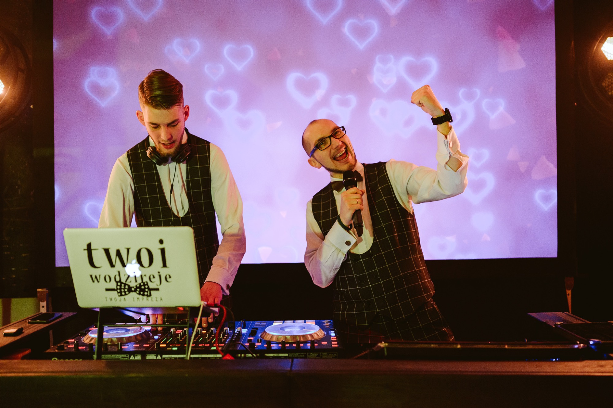 Dwóch wodzirejów w kamizelkach w kratę, jeden z mikrofonem, drugi obsługuje sprzęt DJski z laptopem z logo firmy na tle ekranu z motywem serc.