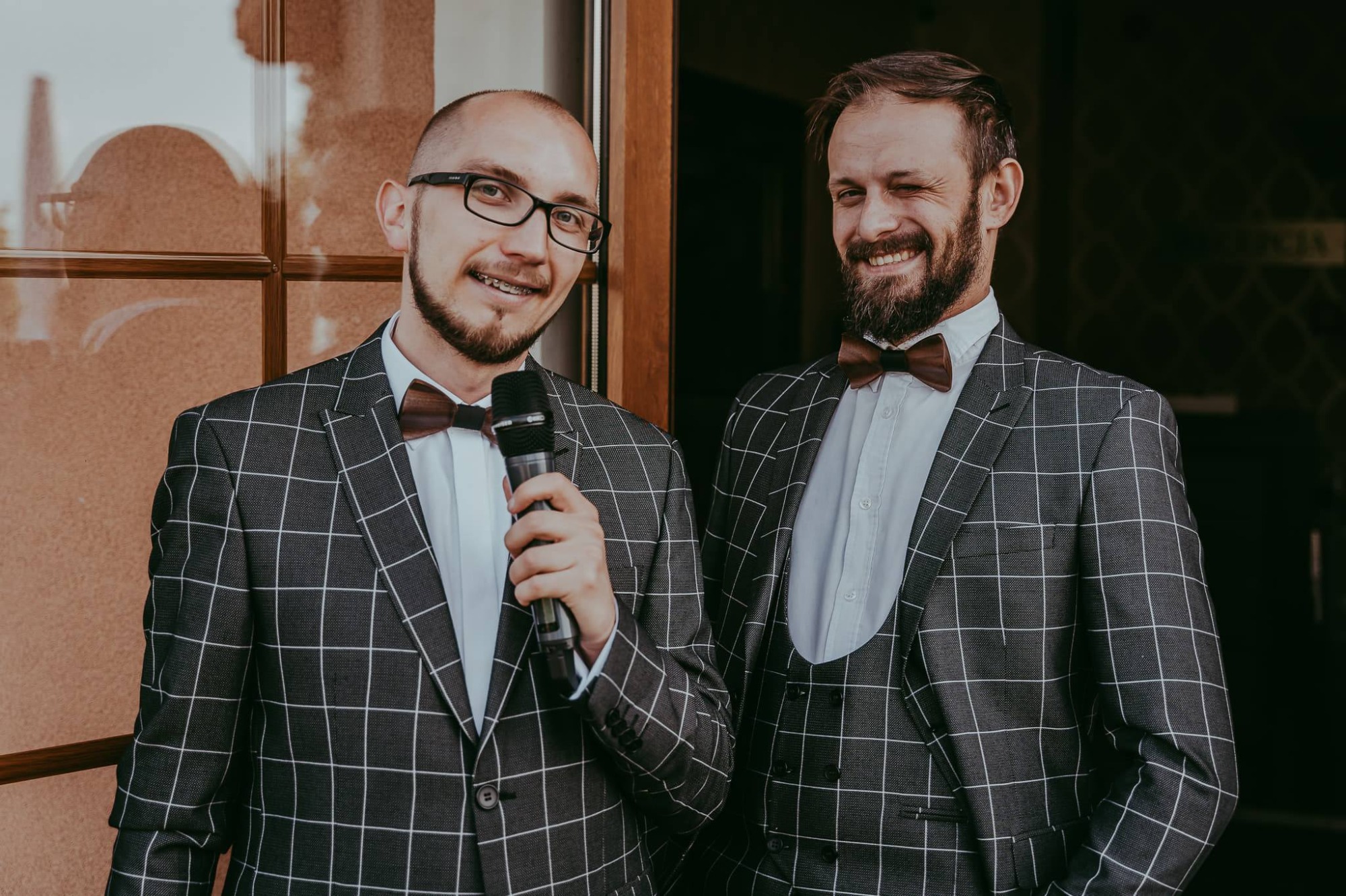 Dwóch mężczyzn w kratkowanych garniturach i drewnianych muchach, jeden z mikrofonem, uśmiechają się do kamery, drugi mruga.