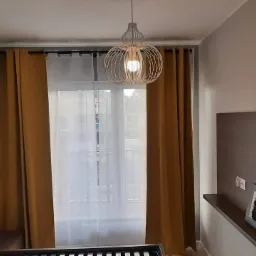 Pokój z oknem przesłoniętym złotymi i białymi zasłonami, nad oknem karnisz, a na suficie ażurowa lampa w kształcie kuli.