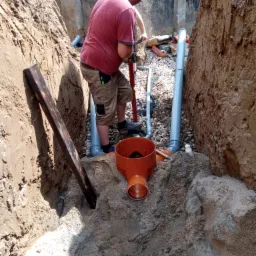 Instalacja kanalizacji: pracownik łączy pomarańczowe rury w wykopie, widoczne szare rury kanalizacyjne, narzędzia, ziemia i ściany wykopu.