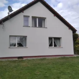 Dom jednorodzinny z nową, białą elewacją w Dźwierzutach, widoczne trzy okna na parterze i dwa na piętrze, antena satelitarna na dachu i fragment drabiny na trawniku.