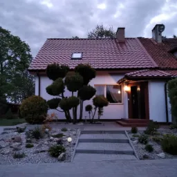 Dwupiętrowy dom z czerwoną dachówką widziany z przodu, z ozdobnymi krzewami i ścieżką prowadzącą do wejścia, oświetlony lampą zewnętrzną, w tle pochmurne niebo.