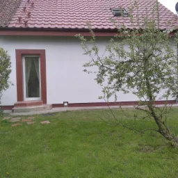 Dom z czerwoną dachówką, ocieplony białą elewacją, z nowym oknem i małym oknem dachowym, widziany z perspektywy trawnika z młodym drzewkiem.