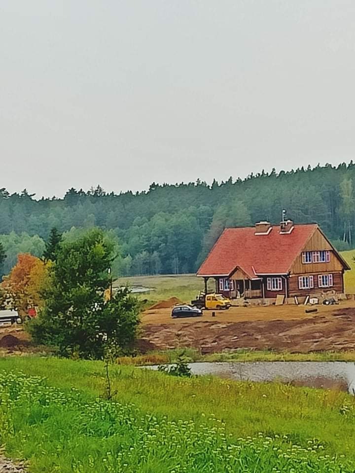 Drewniany dom z czerwoną dachówką w trakcie prac wykończeniowych elewacji, widoczny sprzęt budowlany i przygotowany teren.