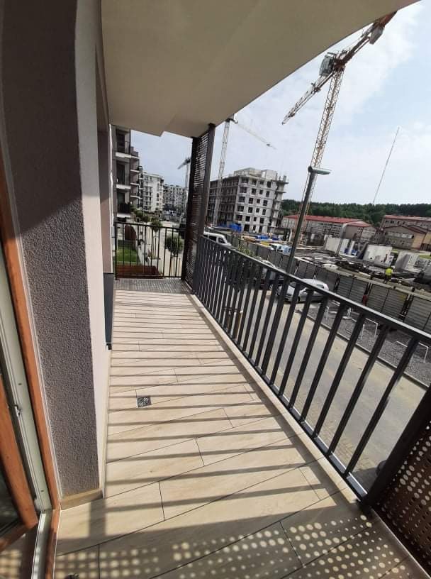 Wykończony balkon z beżową, ułożoną w poprzek terakotą, czarną metalową balustradą i widokiem na plac budowy z żurawiami.